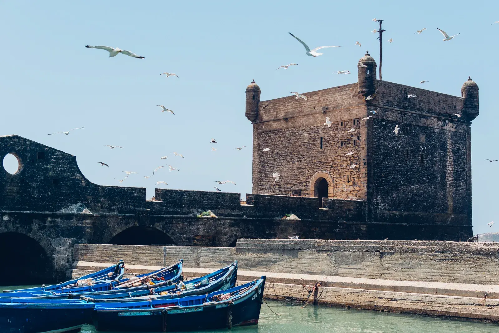 La ciudadela de piedra del puerto de Essaouira, con gaviotas en el cielo y barcas azules abajo