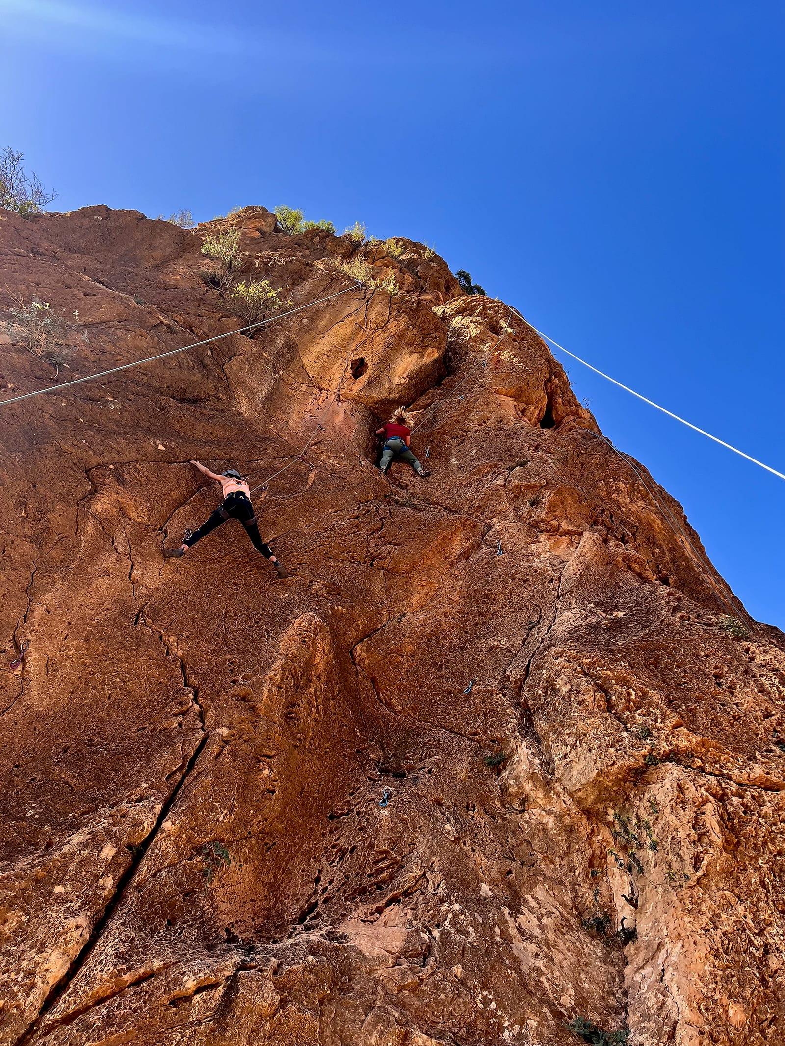 Two guests climbing a red rock face in the Anti-Atlas mountains near Agadir under a deep blue sky